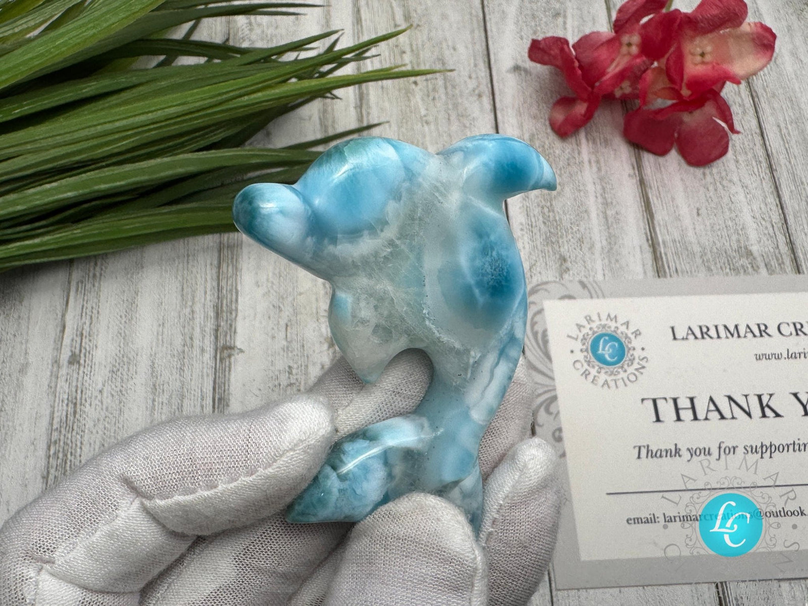 Hand holding a polished larimar dolphin carving with blue and white marbling, displayed on a light wooden surface with