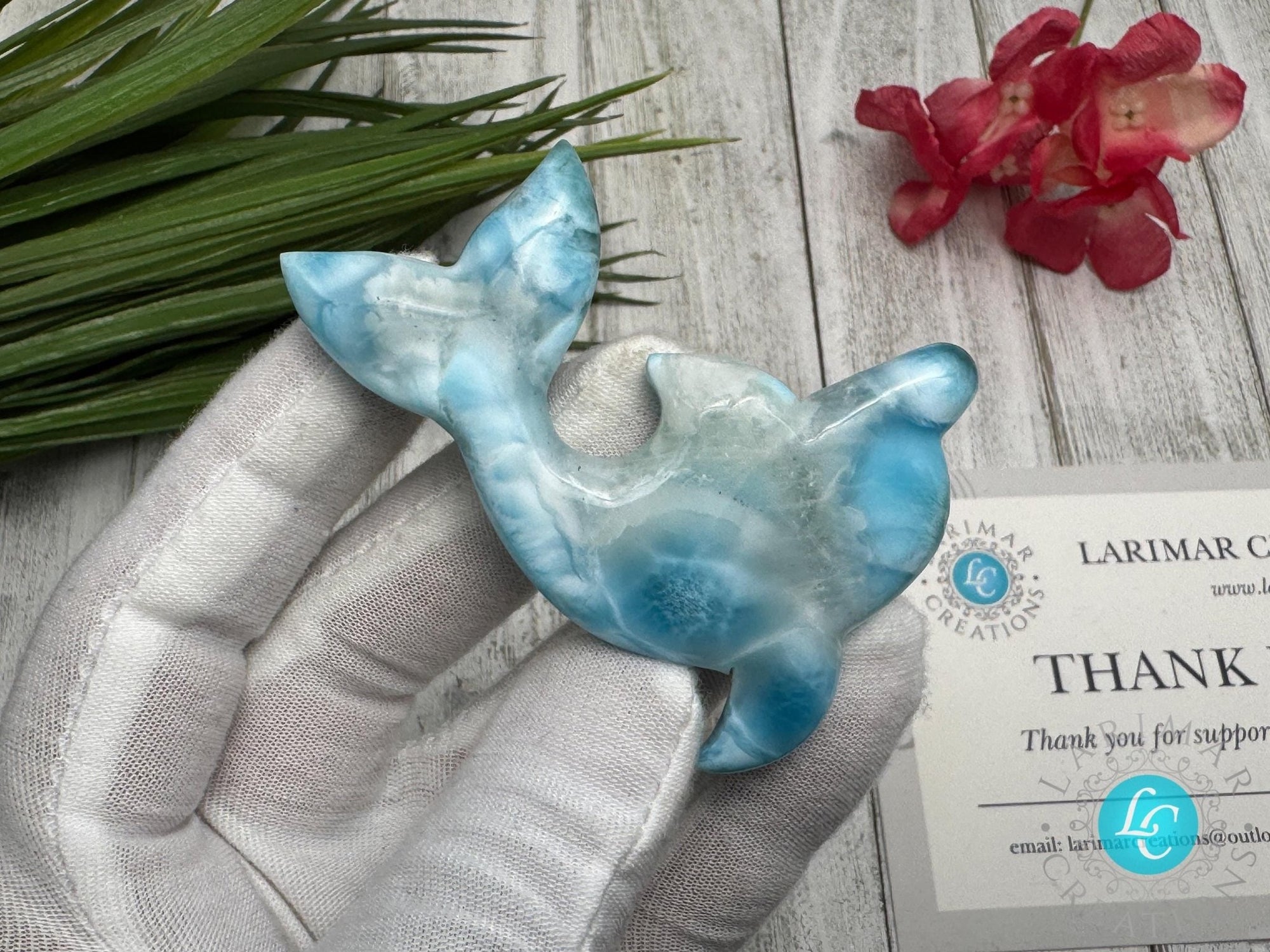 Hand holding a polished larimar dolphin carving with blue and white marbling, displayed on a light wooden surface with
