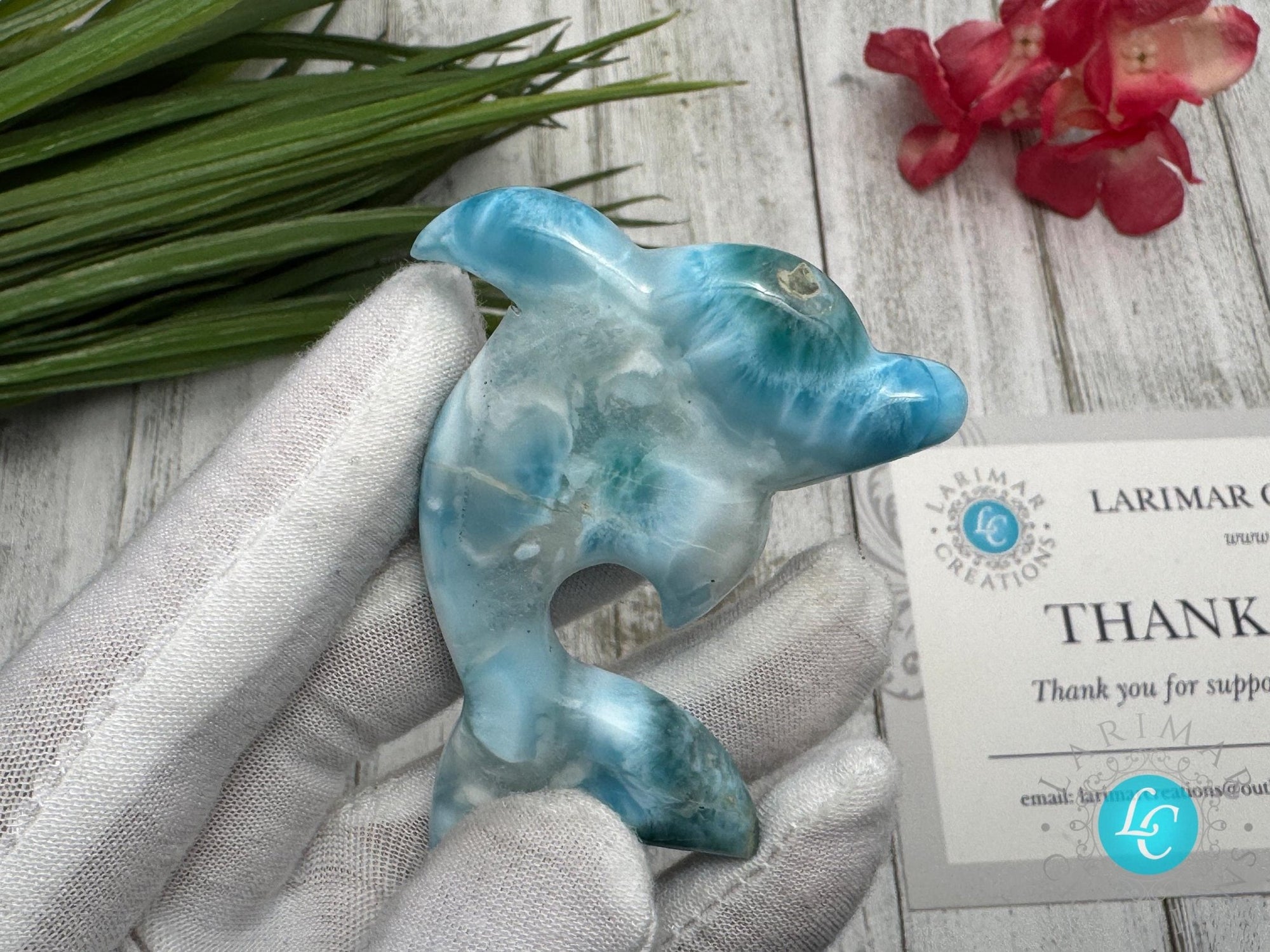 Hand holding a polished larimar dolphin carving with blue and white marbling, displayed on a light wooden surface with