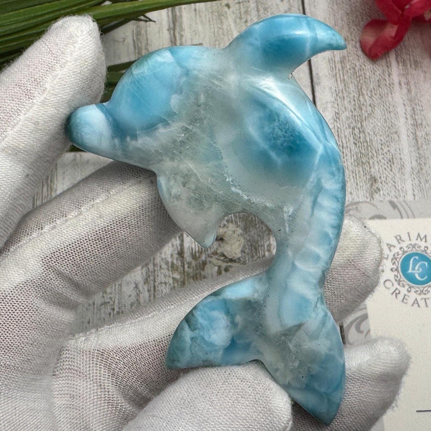 Hand holding a polished larimar dolphin carving with blue and white marbling, displayed on a light wooden surface with