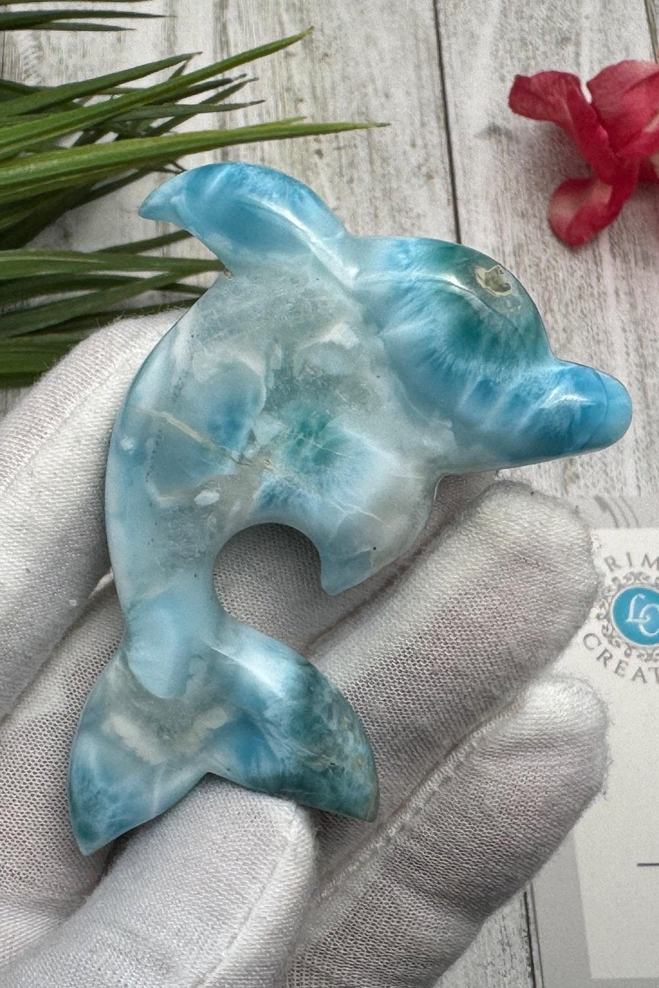 Hand holding a polished larimar dolphin carving with blue and white marbling, displayed on a light wooden surface with