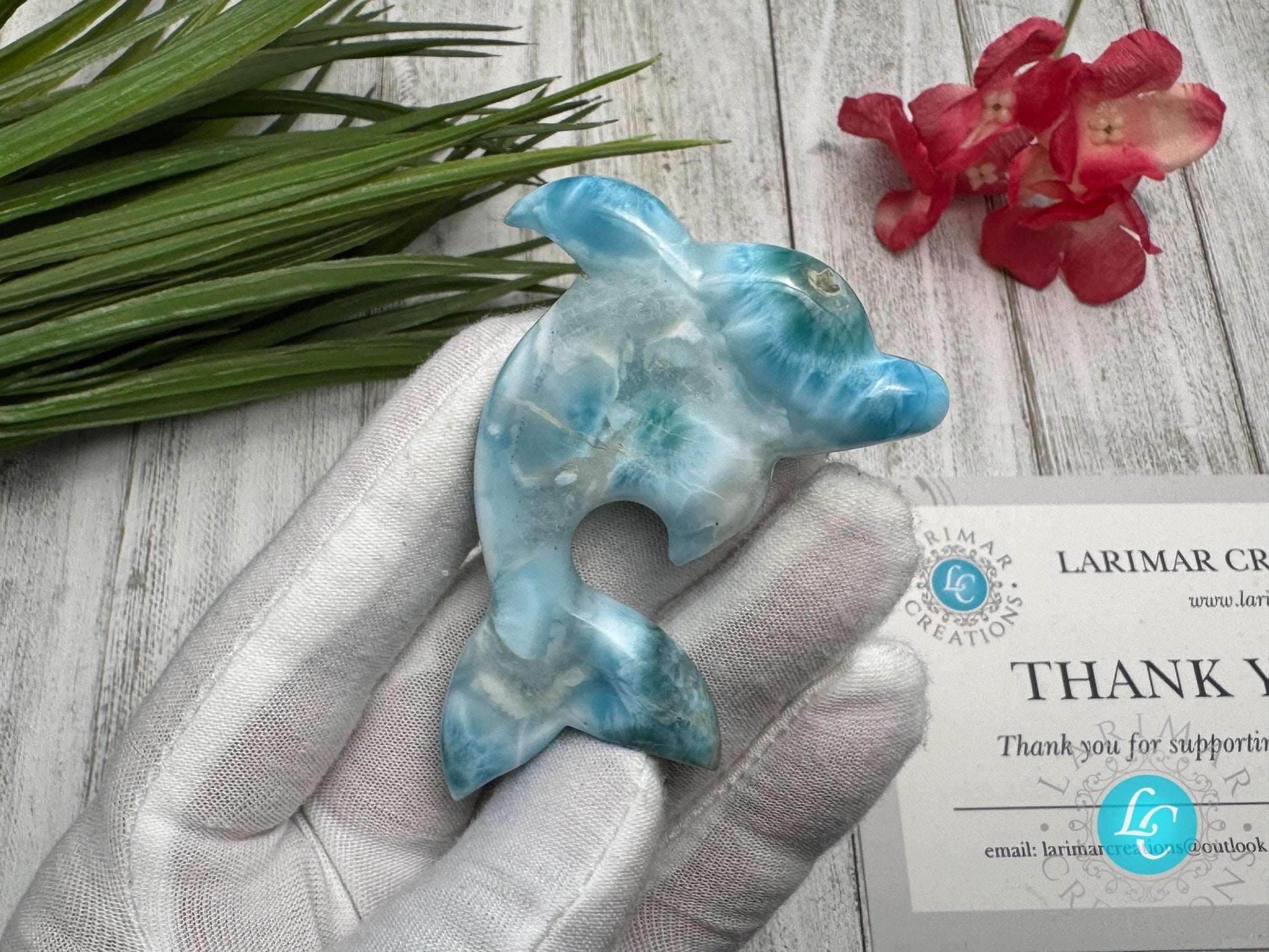 Hand holding a polished larimar dolphin carving with blue and white marbling, displayed on a light wooden surface with