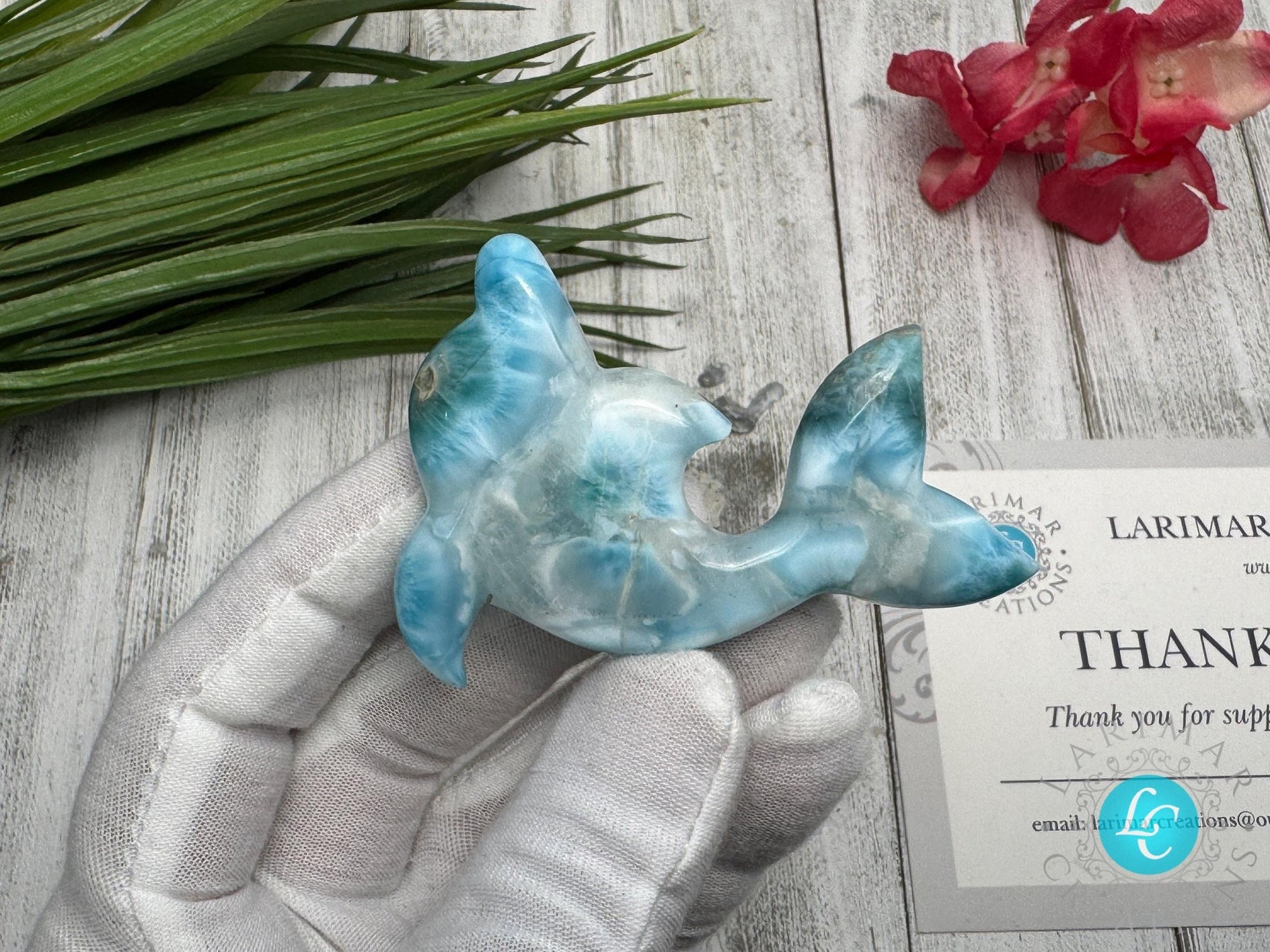 Hand holding a polished larimar dolphin carving with blue and white marbling, displayed on a light wooden surface with
