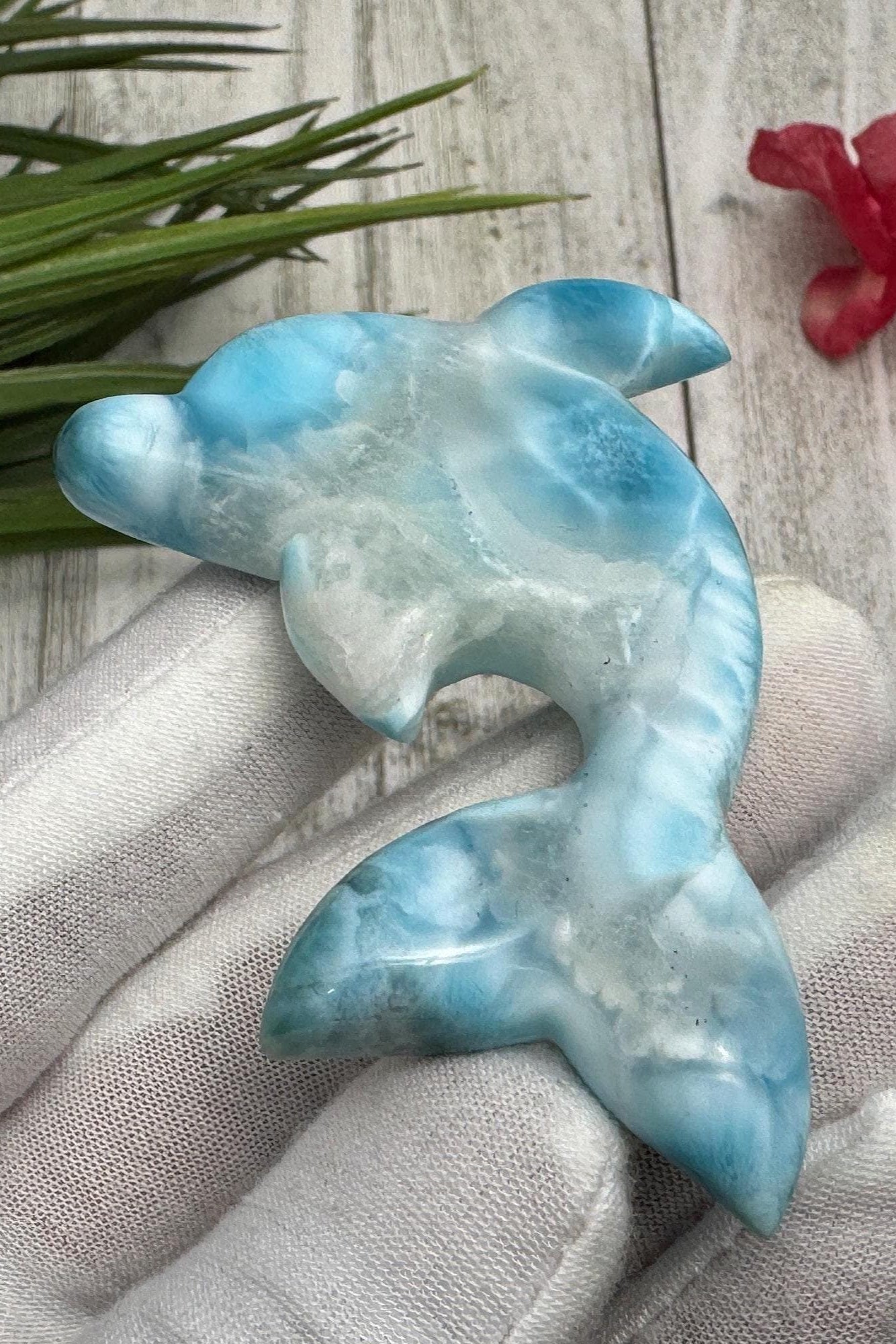 Hand holding a polished larimar dolphin carving with blue and white marbling, displayed on a light wooden surface with