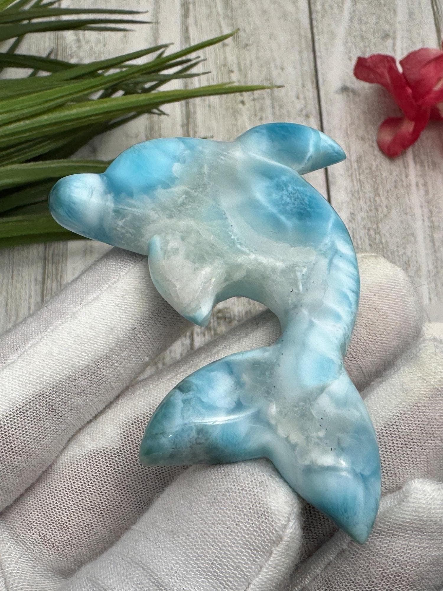 Hand holding a polished larimar dolphin carving with blue and white marbling, displayed on a light wooden surface with