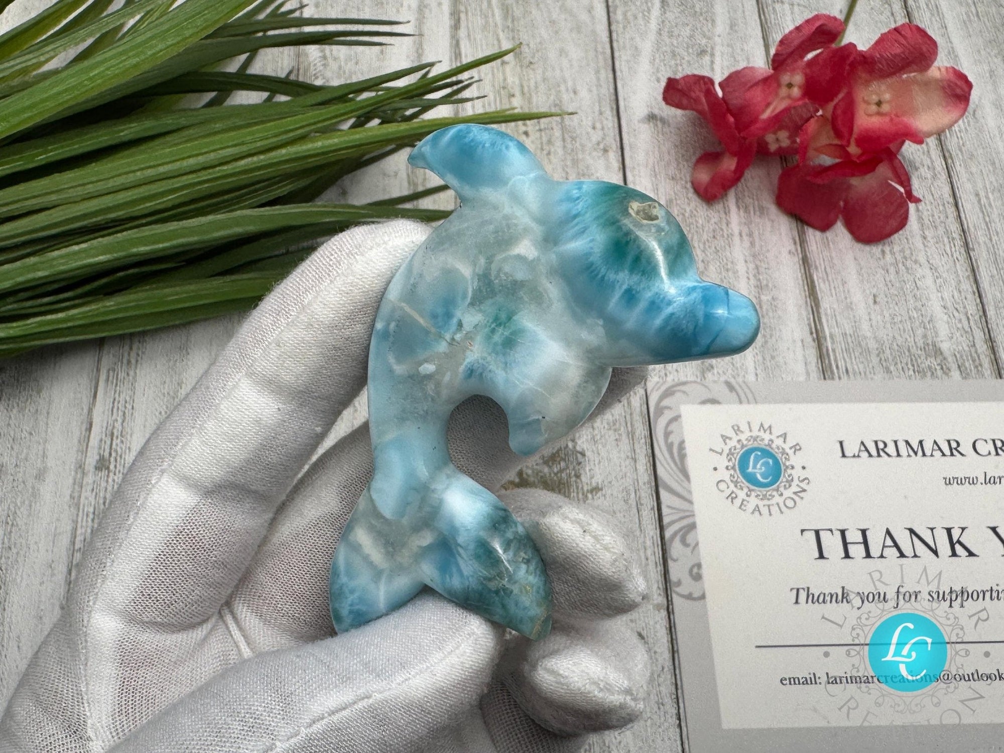 Hand holding a polished larimar dolphin carving with blue and white marbling, displayed on a light wooden surface with