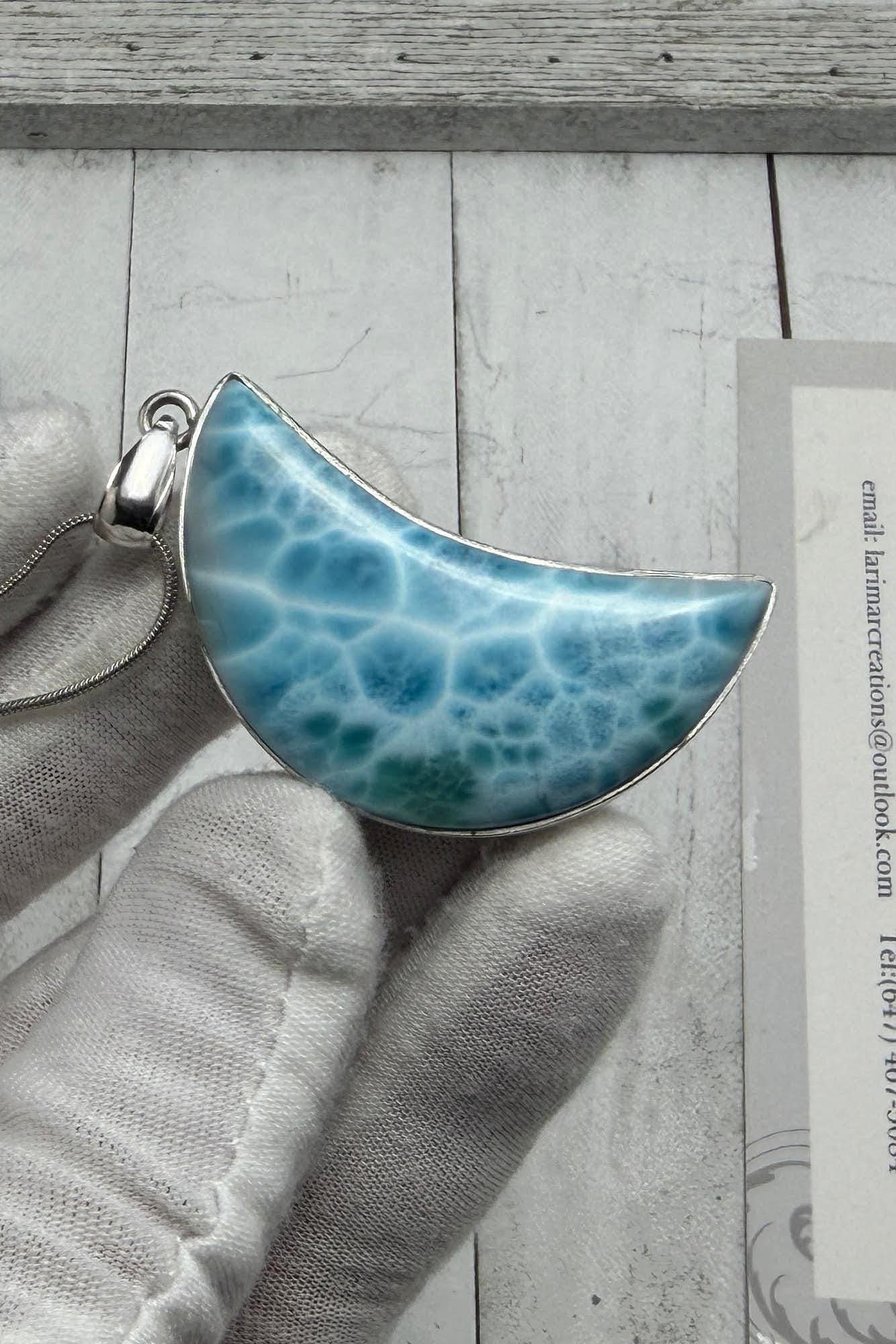 Larimar crescent moon pendant in sterling silver, featuring a natural blue Caribbean gemstone with ocean-wave patterns, held in a gloved hand.