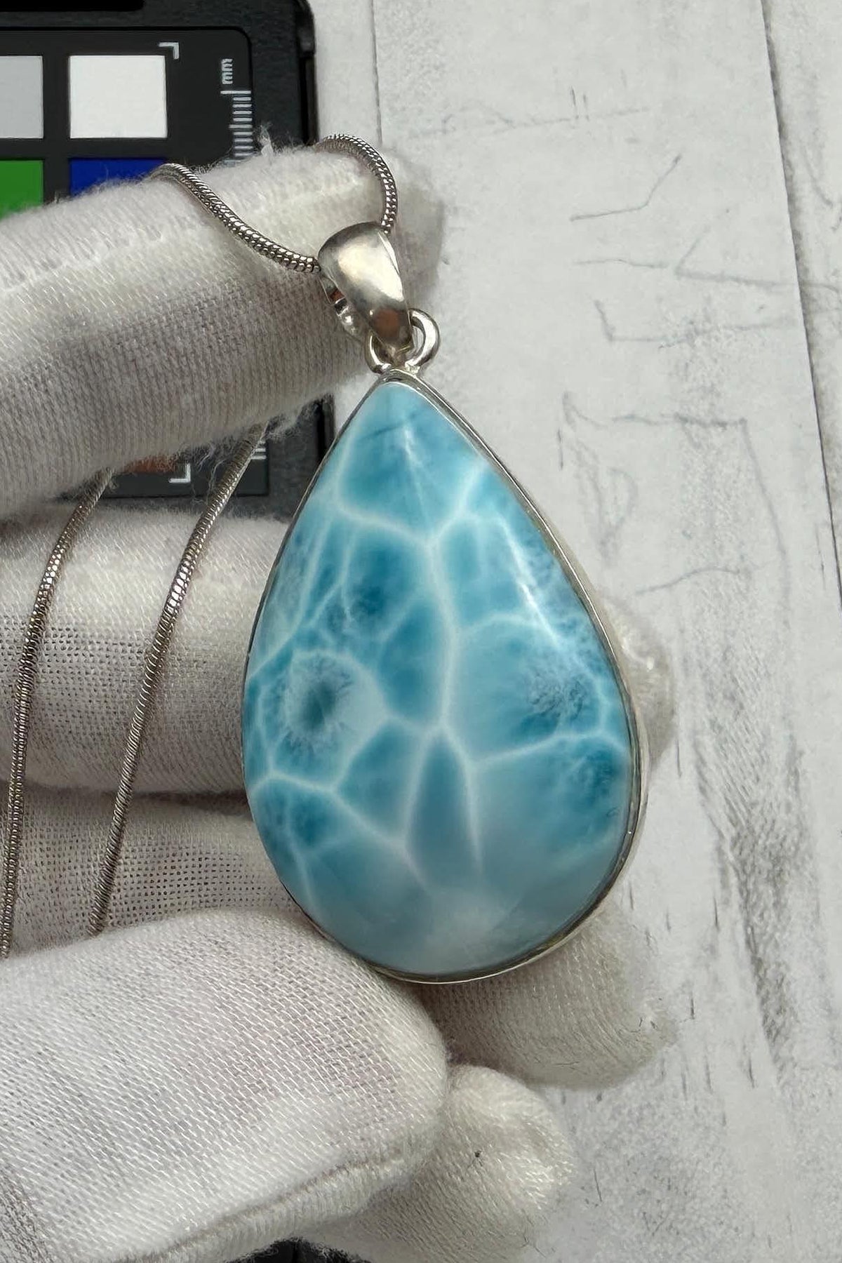 A light blue and white teardrop Larimar pendant necklace in sterling silver, held by a gloved hand.