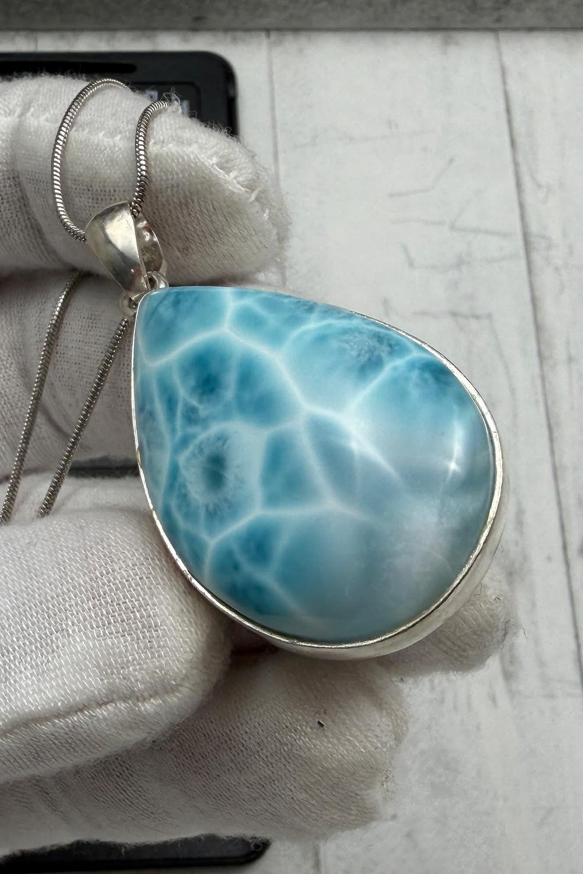A gloved hand holds an authentic teardrop Larimar pendant. The stone features a bright ocean blue color with a white marbled kidney pattern, set in a 925 sterling silver open-back setting on a thin chain.