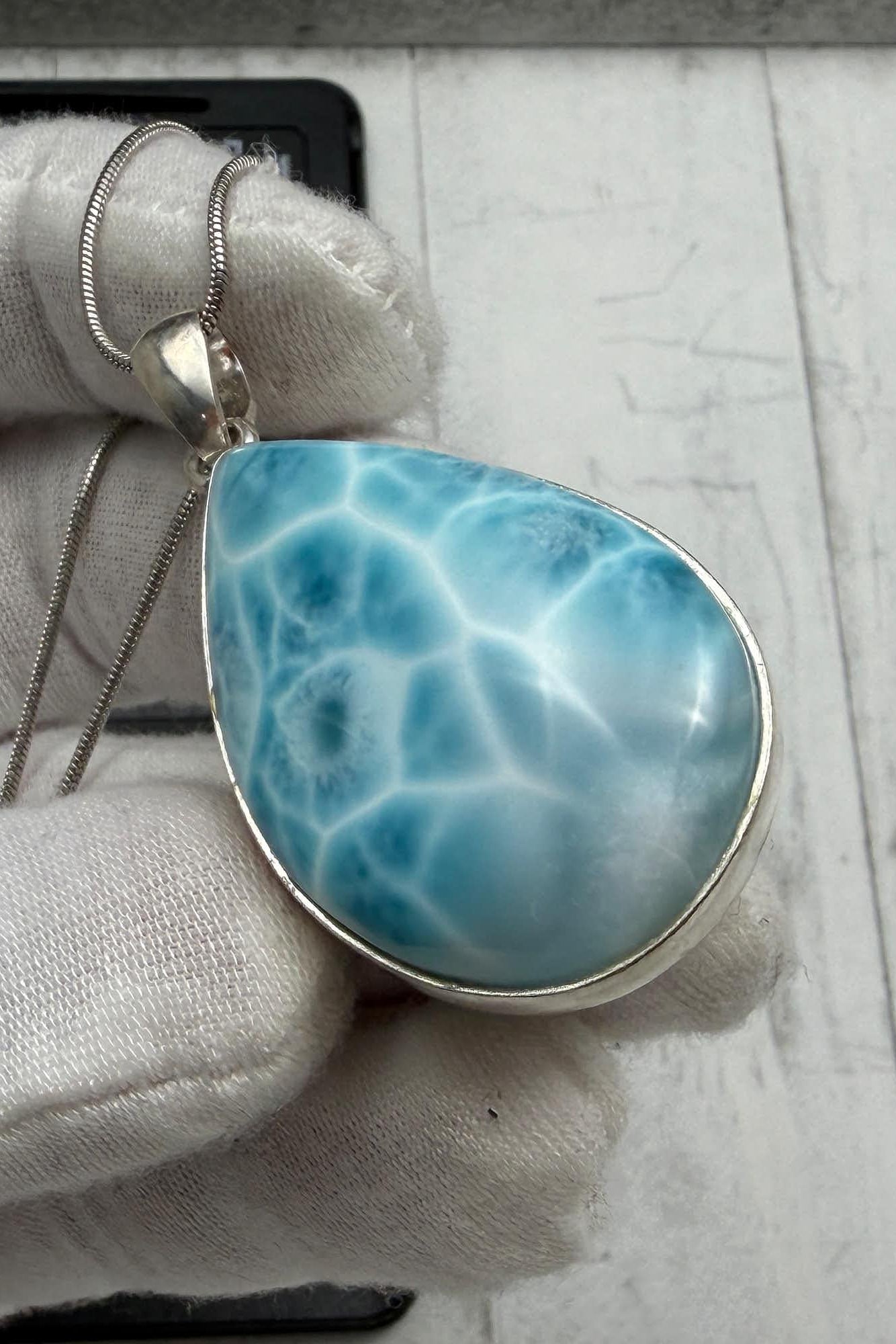 A gloved hand holds an authentic teardrop Larimar pendant. The stone features a bright ocean blue color with a white marbled kidney pattern, set in a 925 sterling silver open-back setting on a thin chain.