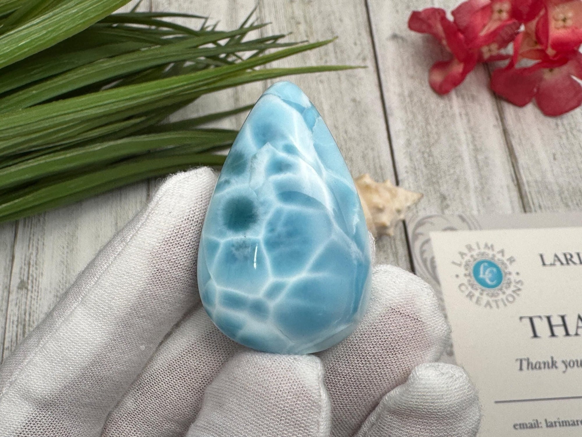 High-quality blue Larimar gemstone with ocean-like patterns, displayed in a white-gloved hand next to a branded