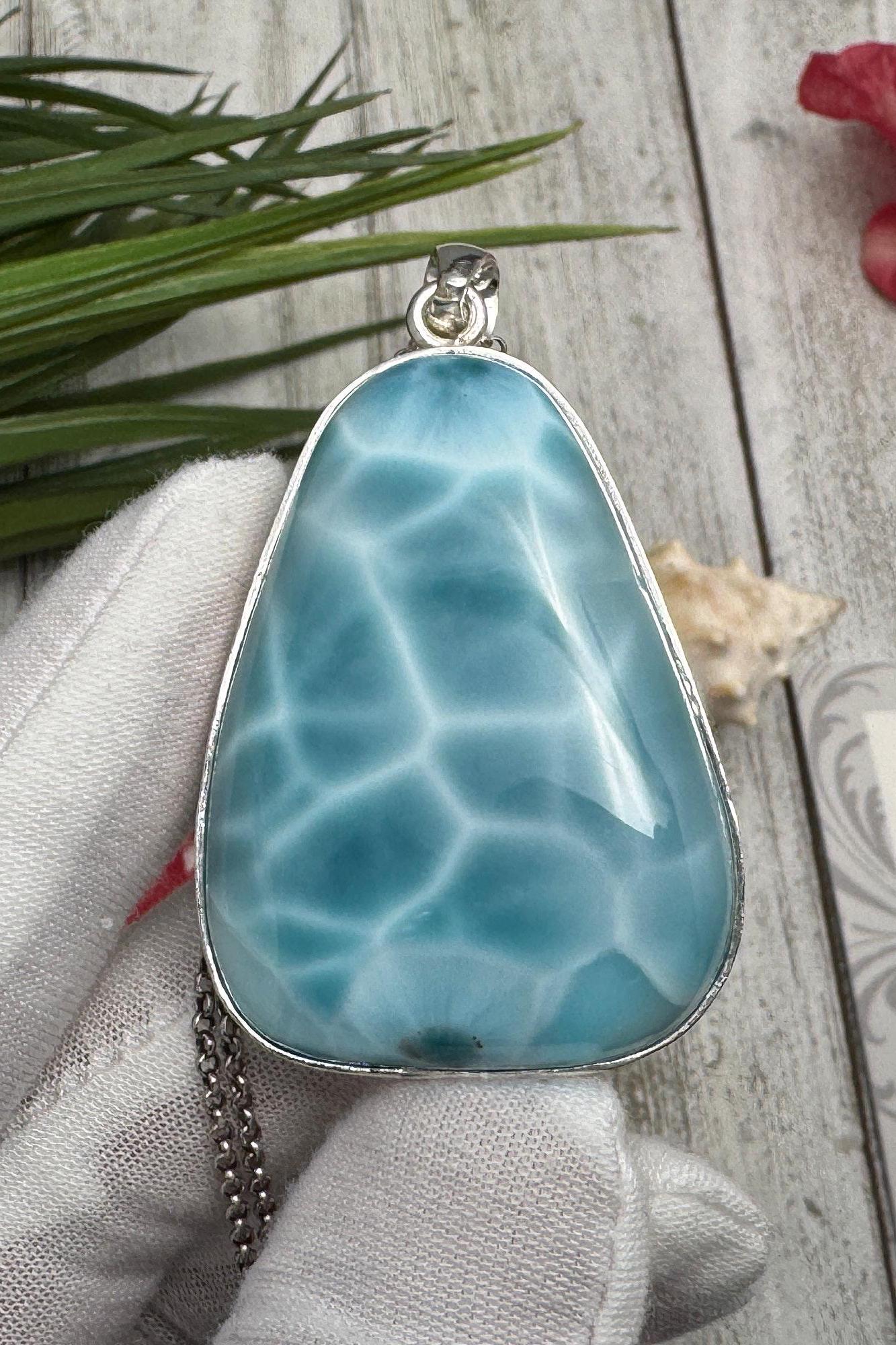 Handcrafted Larimar pendant necklace featuring a natural blue gemstone, held in a white-gloved hand against a rustic