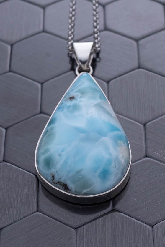 Teardrop-shaped blue pendant on a chain against a hexagonal patterned gray background
