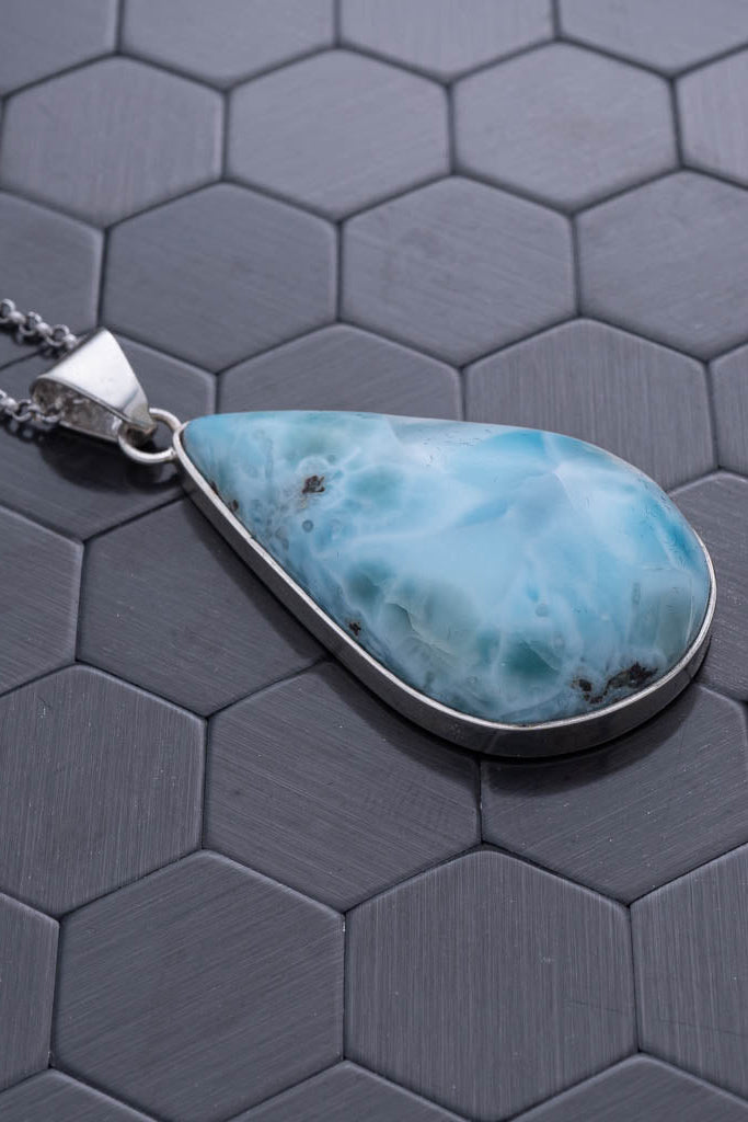 Teardrop-shaped larimar pendant on a silver chain against a hexagonal patterned background