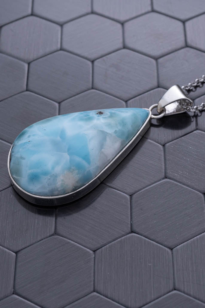 Teardrop-shaped larimar pendant on a chain against a hexagonal patterned background